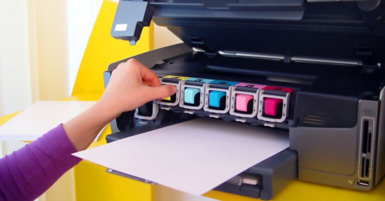 A man's hand inserts a magenta ink cartridge into a printer with cyan, yellow, and black cartridges already in place.