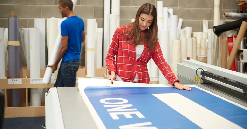 Two individuals working in a print shop, one is inspecting a large printed banner . (Dye Sublimation vs. Digital Printing)